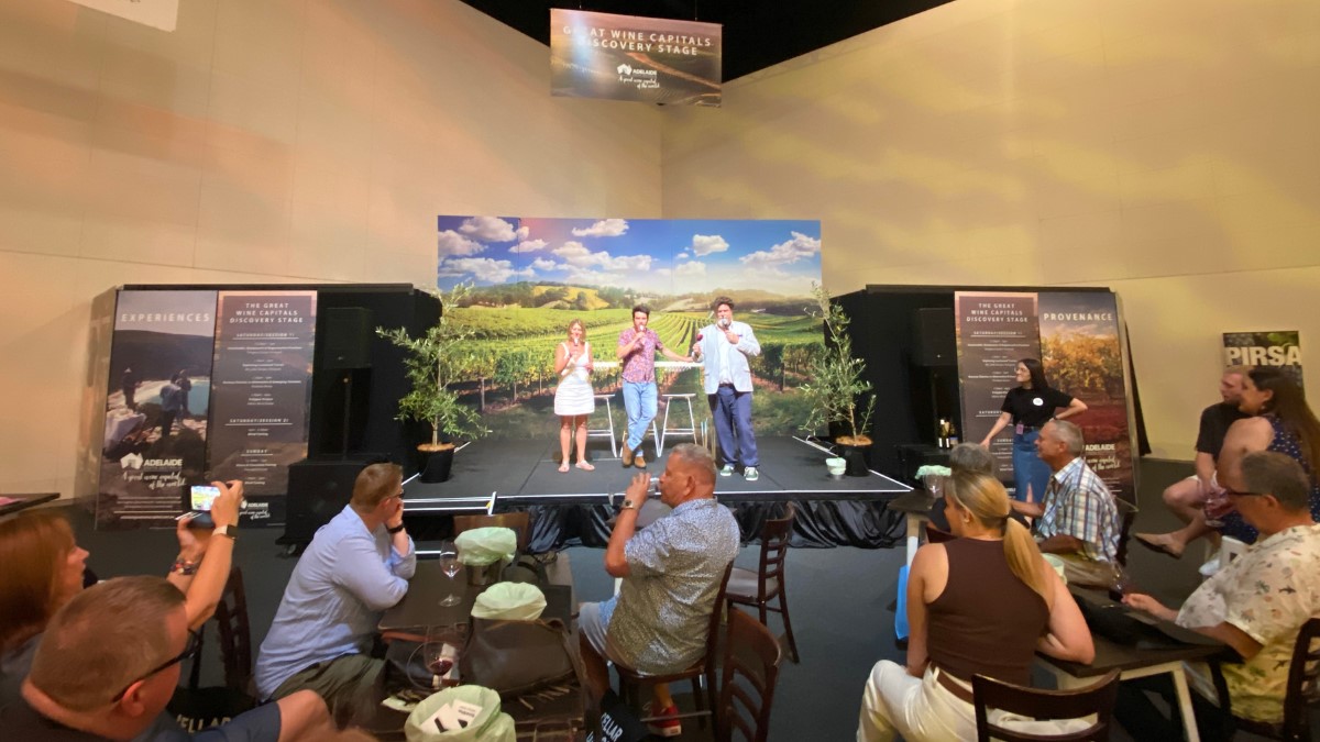 people at tables and chairs looking at 3 people on a stage below a sign saying 'Great Wine Capitals discovery stage